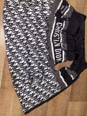 Christian Dior Väst  - Säljer en dunväst från Christian Dior med svartvitt logomönster och tydlig Christian Dior-text på bröstet. Västen har quiltad design och dragkedja framtill. Materialet är syntet och polyester, med en glansig finish på insidan. Perfekt för dig som gillar statement-plagg.