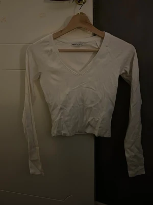 Vit v-ringad långärmad topp Bershka - Säljer en enkel och fräsch vit långärmad topp från Bershka i storlek S. Toppen har en snygg v-ringning och är croppad i modellen, perfekt att matcha med höga jeans eller kjol. Materialet är mjuk bomull som känns skönt mot huden.