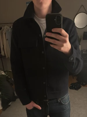 Mörkblå overshirt från New Look - Säljer en stilren mörkblå overshirt från New Look Men i storlek S. Jackan har klassisk krage, stora tryckknappar framtill och en bröstficka. Materialet är ullblandning med slät insida, perfekt för lager-på-lager. Hör bara av dig vid någon fråga.