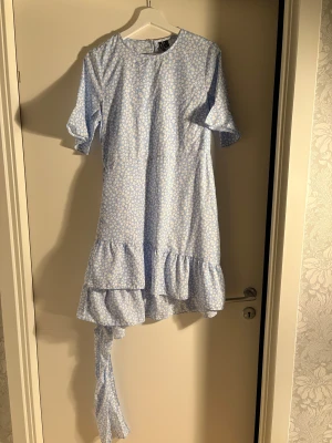 Blommig ljusblå klänning Vero Moda - Supersöt ljusblå klänning från Vero Moda med vitt blommigt mönster. Modellen har korta ärmar, volangdetaljer och asymmetrisk nederdel med extra tygdetalj. Perfekt för dig som gillar romantisk och lekfull stil.