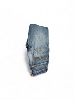 Blå Nudie Jeans  - Säljer ett par blå jeans från Nudie Jeans i storlek W27 L34. Jeansen har klassisk femficksdesign, orange kontrastsömmar och en snygg broderad bakficka. Benen är croppade med rå, fransig kant för en cool och avslappnad vibe. Dom är i 27 34 men avklippta till 27 30! Därav prisen🍾✌️👍