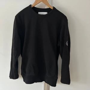 Svart sweatshirt från Calvin Klein Jeans - Svart sweatshirt från Calvin Klein Jeans med klassisk rund hals och ribbade muddar. På ena ärmen sitter en snygg CK-logga i vitt. 