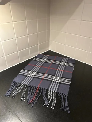 Mörkblå rutig halsduk med fransar - Snygg halsduk i mörkblått med klassiskt rutigt mönster i vitt, svart och röda linjer. Halsduken har fransar i ändarna och är perfekt att slänga över axlarna för en cool look. Materialet känns mjukt och skönt mot huden.liknade burberry