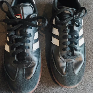 Adidas Samba svart/vit strl 39 1/3 - Klassiska Adidas Samba sneakers i svart skinn och mocka med vita ränder och brun gummisula. Ikonisk retrostil med snörning och rund tå. Perfekta för dig som gillar streetwear och sportig vibe. Det ända att anmärka på är att lite har försvunnit av texten på sulan på insidan. I övrigt i fint skick. 