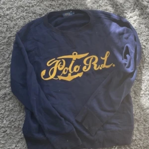 Mörkblå tröja Polo Ralph Lauren XL - Mörkblå långärmad tröja från Polo Ralph Lauren i storlek XL. Framsidan har ett stort gult Polo R.L.-tryck med ankare. Klassisk rund halsringning och ribbade muddar. Perfekt för dig som gillar preppy stil och vill sticka ut med en snygg logga. DEN ÄR XL I BARN