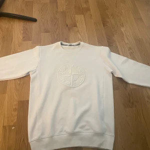 Vit sweatshirt från Stone Island - Vit sweatshirt från Stone Island med stort broderat logomärke på bröstet. Klassisk rund halsringning och långa ärmar. Tröjan har en clean look och är tillverkad i mjukt bomullsmaterial, perfekt för en stilren och avslappnad outfit. storleken är XL men sitter jätte bra på M och L. Tveka inte med att ställa frågor.