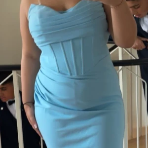 Ljusblå axelbandslös långklänning - Säljer en elegant ljusblå långklänning med axelbandslös design och figurnära passform. Klänningen har draperade detaljer upptill och markerad midja med sömmar som ger en snygg siluett. Perfekt för speciella tillfällen. Använd i endast ett tillfälle i ett par timmar. 