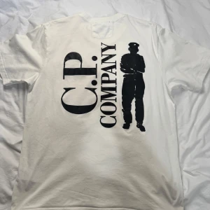 Vit C.P. Company t-shirt med tryck - Säljer en vit t-shirt från C.P. Company med svart logga på bröstet och stort tryck med text och siluett på ryggen. Klassisk rund hals och korta ärmar. Skön och stilren modell i mjuk bomull, perfekt för dig som gillar streetwear med en twist. Storlek M, men passar både M & L
