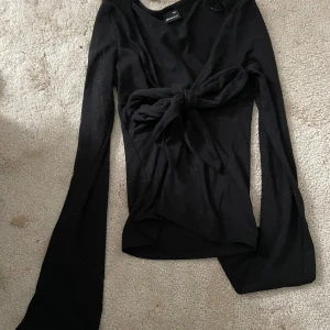 Knyt topp från Gina  - Svart långärmad topp från Gina Tricot i storlek XXS. Modellen har en snygg knytning framtill som ger en cool och trendig look. Toppens material är mjukt och stretchigt, perfekt att styla med jeans eller kjol för en clean vibe.
