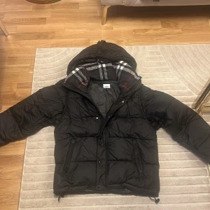 Svart pufferjacka från Burberry med avtagbara ärmar - Svart pufferjacka från Burberry med avtagbara ärmar så du kan bära den som väst. Jackan har huva med rutigt foder i grått, vitt och rött, dragkedjor på fickorna och snörning nertill. Perfekt för kalla dagar när du vill vara både varm och stilren.