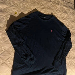 Svart långärmad tröja Polo Ralph Lauren - Svart långärmad tröja från Polo Ralph Lauren i 100% bomull. Klassisk rund halsringning och ikonisk rosa broderad logga på bröstet. Perfekt för en clean och stilren look.