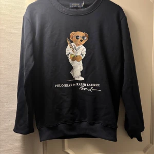 Mörkblå Polo Bear tröja från Ralph Lauren - Mörkblå sweatshirt från Ralph Lauren med ikoniska Polo Bear-trycket på bröstet. Tröjan har rund hals, långa ärmar och ribbade muddar. Tillverkad i mjuk bomull, perfekt för en chill och sportig look.