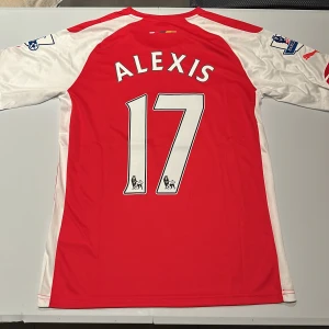 Arsenal Alexis 17 matchtröja Puma - Röd och vit Arsenal fotbollströja från Puma med Alexis och nummer 17 på ryggen. Premier League-patch på ärmarna och Fly Emirates-tryck på bröstet. Klassisk design i lätt och ventilerande material, perfekt för fans av Alexis Sánchez. Replika, endast handtvätt gäller!