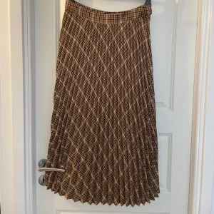 Rutig plisserad kjol från Gina Tricot - Superfin plisserad långkjol från Gina Tricot i brunt och beige rutigt mönster. Kjolen har hög midja och ett klassiskt rutigt print som ger en snygg vintage vibe. Perfekt att styla med chunky boots eller en stickad tröja.