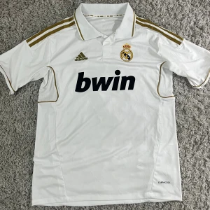 Real Madrid Ronaldo - Vit Real Madrid matchtröja från Adidas med guldiga detaljer, klubbmärke och Bwin tryck på bröstet. På ryggen står det Ronaldo och nummer 7 i guld. Tillverkad i lätt och ventilerande material, den passade inte, darför jag säljer.