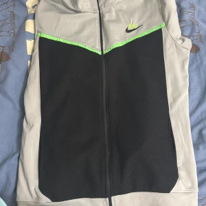 Nike sportväst i svart och grå - Sportig Nike tech  från Nike i svart och ljusgrått med neongröna detaljer och dragkedja framtill. Västen har en modern look med mesh-liknande svart tyg på framsidan och diskret Nike-logga på bröstet. Perfekt för träning eller lager-på-lager.