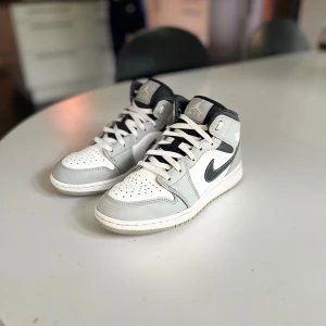 Nike Air Jordan 1 Mid vit/grå - Nike Air Jordan 1 Mid i vitt och ljusgrått med svarta detaljer.  Endast använda ett fåtal gånger men har några skavanker som smutsiga skosnören! Nypris är ca 2000 kr! 