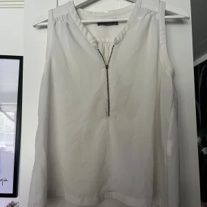 Vit ärmlös blus från Vero Moda XS - Snygg vit blus från Vero Moda i storlek XS. Modellen är ärmlös med en markerad axelparti och har en cool dragkedja framtill. Tyget är lätt och luftigt, perfekt för varmare dagar. Passar dig som gillar stilrena och enkla plagg med en twist.