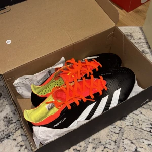 Adidas Predator fotbollsskor svart/orange - Säljer ett par Adidas Predator fotbollsskor i svart med vita detaljer och orange snörning. Skorna har en låg profil, plös i mesh och insida i neongult. Yttersulan har dobbar för gräsplan. Perfekta för dig som vill sticka ut på planen. Nästan helt oanvända, bara testade. Inklusive kvitto