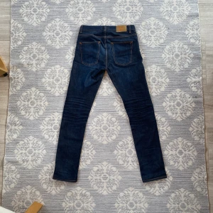 Nudie jeans grim Tim  - Säljer ett par mörkblå slim jeans från Nudie Jeans med klassiska orange sömmar och läderpatch bak. Jeansen är tillverkade i 99% bomull och 1% elastan för en bekväm stretchig känsla. Snygg passform med slimade ben och femficksdesign. Nypris 1699. 
