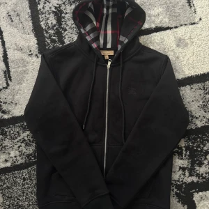 Burberry hoodie - Svart hoodie från Burberry i bomull med klassiskt rutigt foder i huvan. Tröjan har hel dragkedja, snörning i huvan och två fickor framtill. Perfekt för dig som gillar stilren design med en touch av lyx.