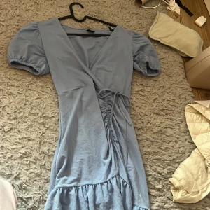 Ljusblå omlottklänning Gina Tricot - Supersöt ljusblå omlottklänning från Gina Tricot i storlek XS. Klänningen har puffiga korta ärmar, knytband i midjan och volang nedtill. Perfekt för dig som gillar en feminin och trendig look. Materialet känns mjukt och skönt mot huden.