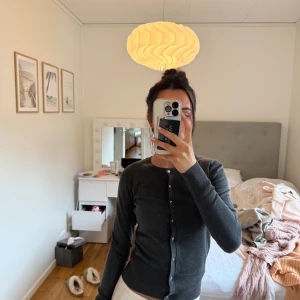 Mörkgrå kofta med knappar - Mörkgrå, klassisk kofta med rund hals och guldiga knappar hela vägen framtill. Långärmad och i mjukt, tunt stickat material som sitter snyggt och har ribbade muddar vid ärmslut och nederkant. Perfekt att slänga över en topp eller bära stängd.