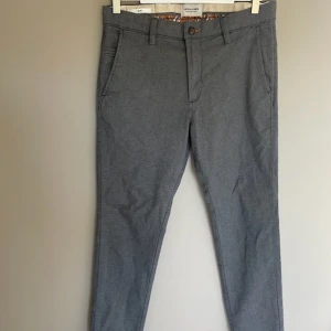 Grå kostymbyxor från Jack & Jones - Slim fit kostymbyxor från Jack & Jones i snygg grå färg med diskret mönster. Byxorna har klassiska fickor fram och bak, bälteshällor och knappstängning. Insidan har en cool detalj med orange och grått mönster. Perfekt för dig som gillar stilrena och moderna vibes.