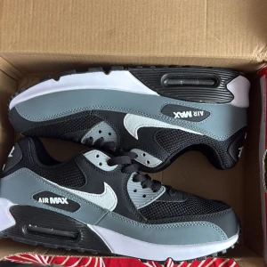 Nike Air Max 90 i svart och grå - Nike Air Max 90 sneakers i svart, grått och vitt. Klassisk siluett med vit swoosh, mesh och syntetmaterial, samt synlig Air Max-dämpning i sulan. Perfekta för dig som gillar streetstyle och sportig look.