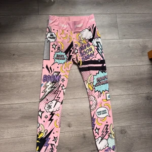 Färgglada leggings med serietidningsmönster - Säljer ett par rosa leggings från STRONGER med coolt serietidningsinspirerat mönster i lila, gult, svart och vitt. De har hög midja och text på linningen. Stolekn är M. Perfekta för dig som vill sticka ut på gymmet eller i vardagen!