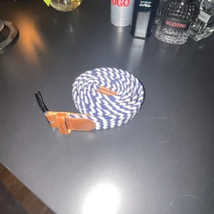 Randigt blåvitt flätat bälte  - Säljer ett snyggt randigt bälte i blått och vitt med bruna läderdetaljer vid spännet. Bältet har en flätad design som ger en cool och modern look. Perfekt accessoar för att lyfta din outfit.