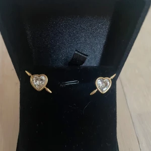 Guldiga hjärtformade örhängen med kristall - Supersöta örhängen i guld med hjärtformad design och glittrande kristall i mitten. Perfekta för dig som vill ha något romantiskt och stilrent.   är riktigt fina att bära till din favoritoutfit. Rostfri 