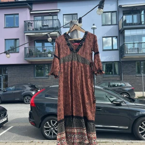Bohemisk klänning Indiska - Snygg bohemisk långklänning i brunt och svart med paisleymönster och kontrasterande detaljer. Klänningen har v-ringning, trekvartsärmar och volang vid ärmslut. Perfekt för dig som gillar en avslappnad och unik stil.
