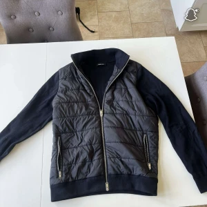 Svart quiltad jacka från Massimo Dutti - Säljer en svart cardigan och den är quiltad jacka från Massimo Dutti med stickade ärmar och krage. Jackan har två fickor med dragkedja och hel dragkedja framtill. Perfekt för dig som vill ha en stilren och bekväm jacka med snygga detaljer.