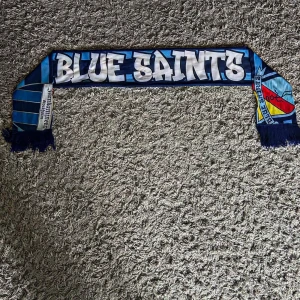 Blå Järnkaminerna halsduk - Snygg supporterhalsduk med texten 'BLUE SAINTS' på ena sidan och 'JÄRNKAMINERNA' på andra. Färgerna är blå, vit och lite röd och gul i klubbmärket. Halsduken har fransar i ändarna och är perfekt för att visa ditt stöd på matchen. Den är skrynklig vid änderna eftersom den har blivit knuten men går att fixa genom att stryka. Priset är inte fastspikat men kommer inte sänka det för mycket.