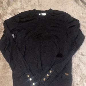 Svart stickad tröja med guldknappar - Svart stickad tröja från Lindex med rund halsringning och långa ärmar. Snygga guldknappar på ärmsluten ger en extra touch. Tröjan har en klassisk passform och diskret metallogo vid höften. Perfekt för dig som gillar stilrena detaljer. KÖPAREN STÅR FÖR FRAKT! 📦 