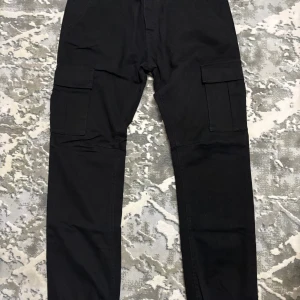 Svarta cargobyxor från Saligia - Svarta cargobyxor från Saligia med raka ben och stora fickor på sidorna. Byxorna har klassisk cargostil med bälteshällor och knappstängning. Perfekta för en avslappnad och cool look.