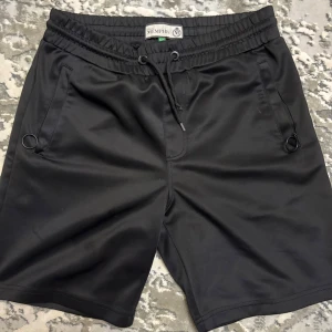 Svarta shorts från Memphis med snörning - Svarta shorts från Memphis i medium med elastisk midja och snörning framtill. Tillverkade i mjukt syntetmaterial, har två sidofickor med ringdetaljer och en bakficka med mönstrad kant. Perfekta för chill eller träning.