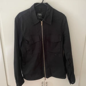 Svart overshirt från Zara med dragkedja - Stilren svart overshirt från Zara med raka linjer, två stora bröstfickor och silverfärgad dragkedja framtill. Jackan har klassisk krage och är tillverkad i ett mjukt bomullstyg. Perfekt för lager-på-lager och enkel att matcha. ✅                                              Nypris: 799kr mitt pris: 399kr😃✅                           Ställ gärna frågor vid funderingar! ✅