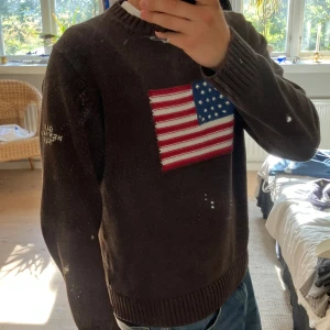 Brun stickad  tröja med USA flagga GANT - Brun stickad tröja från GANT med broderad amerikansk flagga på bröstet och vit text på ena ärmen. Tröjan har rund hals, ribbade muddar och lång ärm. Perfekt för dig som gillar klassisk college-stil och snygga detaljer. Storlek L men sitter som en M. Model är 180 lång.