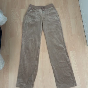 Beiga mjukisbyxor Juicy Couture XS - Supermjuka beiga mjukisbyxor från Juicy Couture i velour med broderad logga framtill, bred resår och snörning i midjan. Byxorna har raka ben och stora fickor både fram och bak med knappdetaljer. Perfekta för en chill och trendig look.
