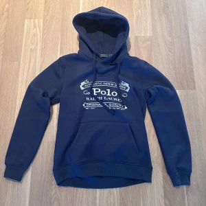 Polo Ralph Lauren hoodie Marinblå - Snygg marinblå hoodie från Polo Ralph Lauren med vit tryckt logga på bröstet. Klassisk känguruficka framtill, huva med snörning och ribbade muddar. Perfekt för en chill och stilren look.