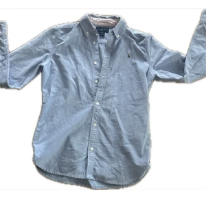 Blå skjorta från Ralph Lauren - Snygg ljusblå skjorta från Ralph Lauren med klassisk krage och knappar framtill. Broderad logga på bröstet och långärmad modell. Tillverkad i mjuk bomull, perfekt för en chill och stilren look. Nypris är runt 1500 kr. Skriv om ni har frågor. Pris kan sänkas. Använd två gånger. Str runt 158 cm.