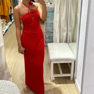 Röd virkad långklänning med blommor - Säljer en röd virkad långklänning med halterneck och dekorativa virkade blommor. Klänningen har en öppen rygg och smickrande passform, perfekt för sommaren. Snygg detalj med knyt i nacken och liten öppning framtill.