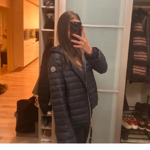 Mörkblå Moncler med huva - Säljer en mörkblå från Moncler med tunn huva och dragkedja framtill. Jackan har ett quiltat mönster och Moncler-logga på ärmen. Perfekt för regninga dagar samt hösten, och snygg passform. Jackan är lätt och smidig att bära. Säljer denna då den inte används, den är i jättebra skick då den knappt har använts. Det är en gammal modell.