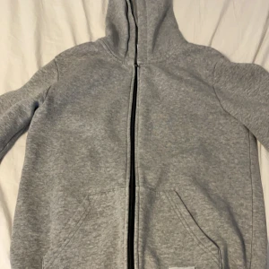 Grå hoodie från Andy by Frank Dandy - Grå hoodie från Andy by Frank Dandy med dragkedja framtill och två stora fickor. Tröjan har huva och ribbade muddar vid ärmslut och nederkant. Perfekt för chill dagar eller när du vill ha en enkel och stilren look.