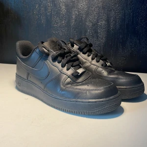 Nike Air Force 1 Low Svarta Sneakers - Svarta Nike Air Force 1 sneakers med klassisk låg siluett och platt sula. Skorna har snörning med metallbricka märkt 'AF1', rund tå och är tillverkade i skinn med textilfoder. Swoosh-logga på sidan och Nike Air broderat på hälen. Perfekt för dig som gillar stilrena och ikoniska sneakers.Pris kan diskuteras!