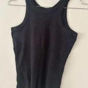 Svart ribbad linnetopp - En svart ribbad linnetopp med klassisk rund halsringning och breda axelband. Toppen är ärmlös och har en tight passform som framhäver siluetten. 