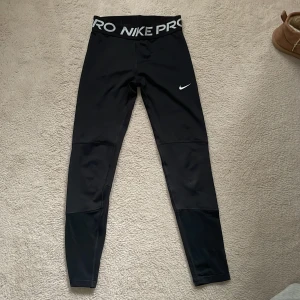 Nike Pro svarta leggings Dri-FIT - Svarta Nike Pro leggings med bred resår i midjan och stor logga. Tillverkade i Dri-FIT-material som andas och transporterar bort fukt. Tight passform och diskret Nike Swoosh på benet. Perfekta för träning eller sport.Tråden har åkt av lite längst ner vid benet men hoppas det inte gör så mycket. Säljer då dom är för små.❣️