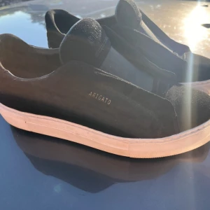 Svarta sneakers från Axel Arigato - Snygga svarta sneakers från Axel Arigato med vit sula. Skorna har elastiska paneler istället för snörning och är i mocka. Perfekta för dig som gillar stilrena och trendiga skor. Mycket bra skick då de bara är använda några få gånger! Storlek 40!